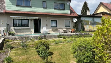 Hausfassade mit Gartenbereich und kleiner Terrasse im Vordergrund unter bewölktem Himmel.