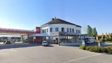 Außenansicht eines Gebäudes mit Tankstelle und Bistro, umgeben von einem großen Parkplatz unter blauem Himmel.