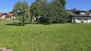 Große Rasenfläche mit Bäumen und einem Wohnhaus im Hintergrund unter blauem Himmel.