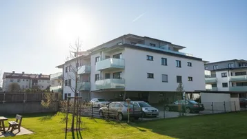 Modernes Mehrfamilienhaus mit Balkonen, Parkplätzen und Grünflächen unter strahlendem Himmel.