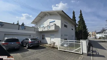 Weißes Einfamilienhaus mit gepflasterter Auffahrt, Garage und zwei geparkten Autos unter blauem Himmel.