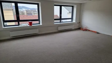 Geräumiger, leerer Büroraum mit Teppichboden, großen Fenstern und Blick auf die Stadt.