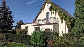 Hausfassade mit Balkon und begrüntem Vorgarten unter blauem Himmel.