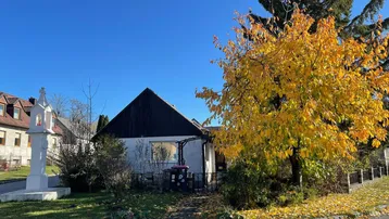 Kleines Haus mit Satteldach und einem Kreuz vor dem Eingangsbereich, umgeben von Herbstbäumen.