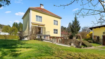 Gelbes Einfamilienhaus mit rotem Dach und großem Garten an einem sonnigen Tag.