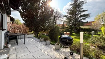 Sonnige Terrasse mit Gartenmöbeln und Grill, umgeben von viel Grün und Bäumen.