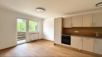 Heller offener Wohnbereich mit integrierter Küchenzeile, Holzboden und Zugang zum Balkon.