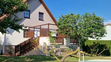 Hausfassade mit großem Garten, kleinem Pool und erhöhter Holzterrasse mit Zugang zum Garten.