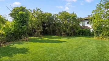 Große Rasenfläche mit üppiger Vegetation und einem Haus im Hintergrund unter blauem Himmel.