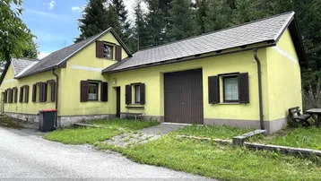 Gelbes Haus mit braunen Fensterläden und Satteldach, umgeben von Bäumen und Wiese.
