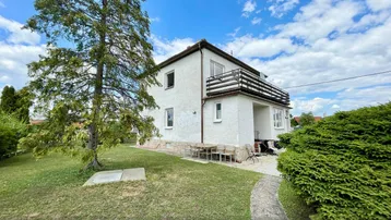 Weißes Einfamilienhaus mit großem Garten, Rasenfläche und Außensitzbereich unter blauem Himmel.
