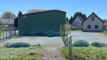Grünes Lagergebäude auf einem Schotterplatz mit umliegenden Wohnhäusern unter blauem Himmel.