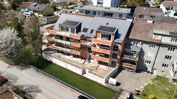 Luftaufnahme eines Neubaus mit roten Ziegelwänden, grauen Dächern und Solarpaneelen am Flussufer.