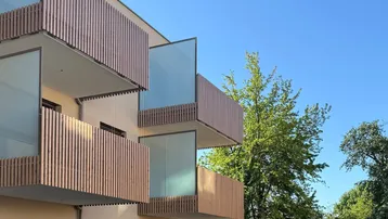 Moderne Gebäudefassade mit gestaffelten Balkonen, Holzlamellen und Glasgeländern unter blauem Himmel, umgeben von Grün.
