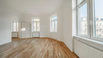 Helles, leeres Zimmer mit Parkettboden und großen Fenstern, die viel Tageslicht hereinlassen.