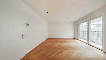 Helles, leeres Wohnzimmer mit modernem Fischgrätparkett und Zugang zu einem Balkon.