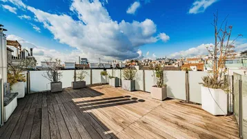 Große Dachterrasse mit Holzboden, Pflanzenkübeln und Stadtblick unter blauem Himmel.