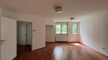 Heller Wohnraum mit warmem Parkettboden, weißen Wänden und Zugang zu einem Außenbereich.