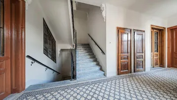 Historisches Treppenhaus mit schmiedeeisernem Geländer, gemusterten Fliesen und Holztüren.