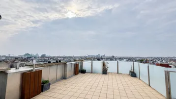 Geräumige Dachterrasse mit Fliesenboden und weitem Blick über die Stadt unter bewölktem Himmel.