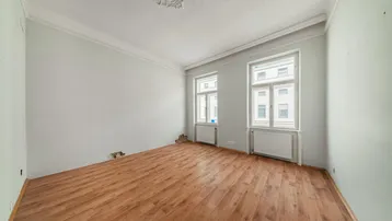 Geräumiges Zimmer mit hellem Laminatboden, zwei Fenstern und Stuckverzierung an der Decke.