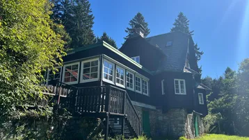 Charmantes Holzhaus mit Balkon und Treppe, umgeben von üppiger Natur und Bäumen.