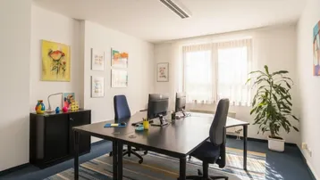 Helles und modernes Büro mit mehreren Arbeitsplätzen, großen Fenstern und Teppichboden.