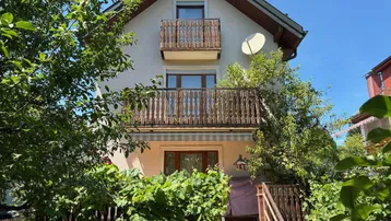 Vorderansicht des Hauses mit zwei Holzbalkonen und üppiger Vegetation im Vorgarten.
