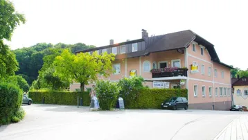 Das Gebäude präsentiert sich mit einer hellrosa Fassade, umgeben von grünen Hecken und Bäumen, an einer ruhigen Straße gelegen.