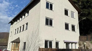 Mehrstöckiges Wohngebäude mit heller Fassade und Fenstern, umgeben von Bäumen und einem Holzzaun.