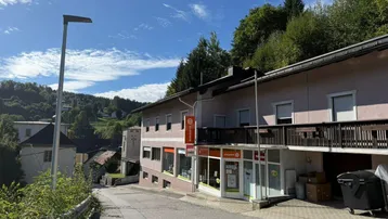 Gebäudeansicht mit Geschäften im Erdgeschoss und Wohnungen darüber, an einer Hangstraße gelegen.
