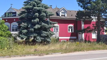 Rotes Gebäude mit weißer Fassade, Satteldach und großen Bäumen an einer Straße gelegen.