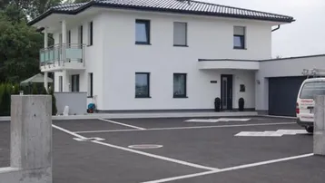 Modernes weißes Einfamilienhaus mit dunklem Dach, Garage und gepflasterter Einfahrt mit Parkplätzen.