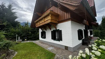 Traditionelles Haus mit Holzbalkon, weißer Fassade und gepflegtem Vorgarten unter bewölktem Himmel.