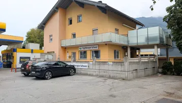 Gelbes Gebäude mit Parkplätzen davor, einer Tankstelle und Bergen im Hintergrund.