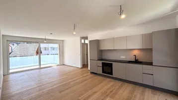 Heller offener Wohnbereich mit moderner Küchenzeile, Holzboden und Zugang zu einem Balkon.