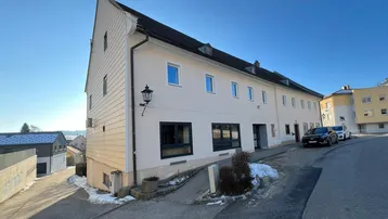 Gebäudeansicht von der Straße mit schräger Zufahrt und heller Fassade unter blauem Himmel.