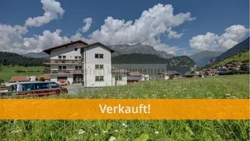 Gebäude in alpiner Landschaft mit grünen Wiesen und Bergen im Hintergrund, mit einem 'Verkauft!'-Banner.