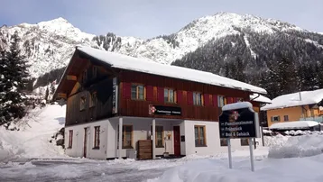 Traditionelles Gebäude mit Holzelementen und roten Fensterläden, umgeben von schneebedeckten Bergen.
