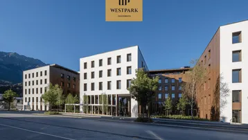 Modernes Gebäude mit weißer und Holzfassade, umgeben von Bäumen und Bergen im Hintergrund unter blauem Himmel.