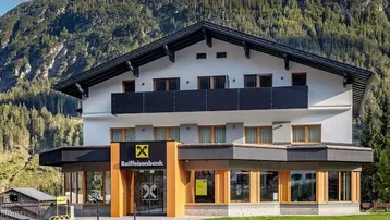 Modernes Gebäude mit Balkonen und großen Fenstern, umgeben von einer Berglandschaft.