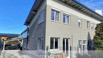 Seitenansicht des Gebäudes mit einer kleinen Terrasse und einem Carport im Hintergrund unter blauem Himmel.