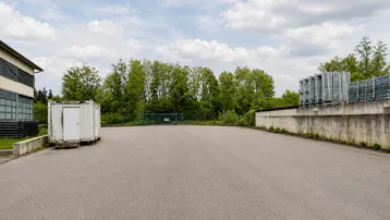 Großer asphaltierter Außenbereich mit einem Gebäude, einem Lagercontainer und Bäumen im Hintergrund unter bewölktem Himmel.