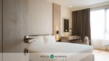 Modernes Schlafzimmer mit großem Bett, hellem Holzdekor und einem Schreibtischbereich. Große Fenster mit braunen Vorhängen sorgen für viel Tageslicht.
