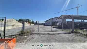 Eingangsbereich eines umzäunten Grundstücks mit Schotterfläche, einem Gebäude und einem Baukran im Hintergrund.