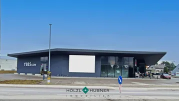Moderne Geschäftsfassade mit großen Glasflächen und Parkplätzen davor unter blauem Himmel.