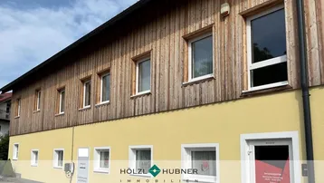 Gebäudefassade mit gelbem Putz im Erdgeschoss und Holzverkleidung im Obergeschoss unter blauem Himmel.