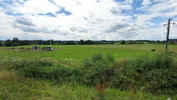 Weite grüne Landschaft mit Feldern und einem kleinen Gebäude unter bewölktem Himmel, typisch für ländliche Gegenden.