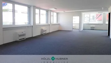 Geräumiger, leerer Büroraum mit Teppichboden und großen Fenstern, die viel Tageslicht hereinlassen.