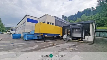 Außenansicht eines Industriegebäudes mit Laderampen, einem gelben Anhänger und Müllcontainern.